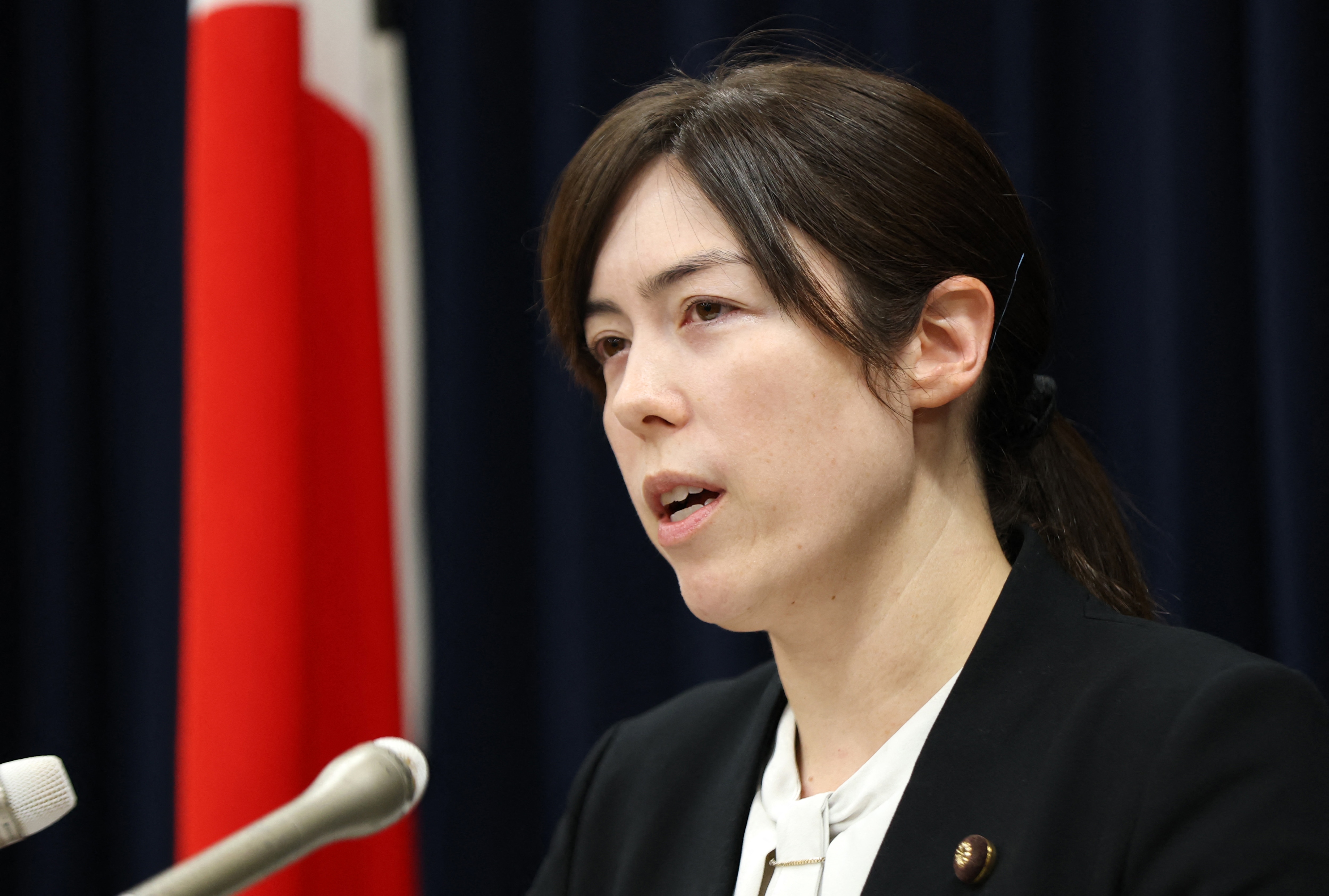 A woman with dark hair and in a dark jacket speaks at a microphone next to a Japanese flag.