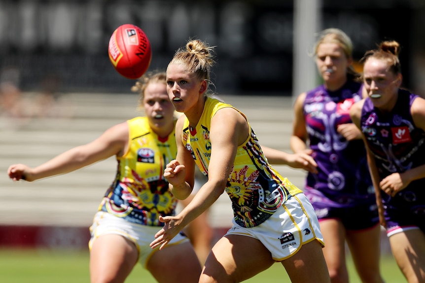 An AFLW player clenches her fist after handballing the ball ahead of her while defenders trail behind her.  