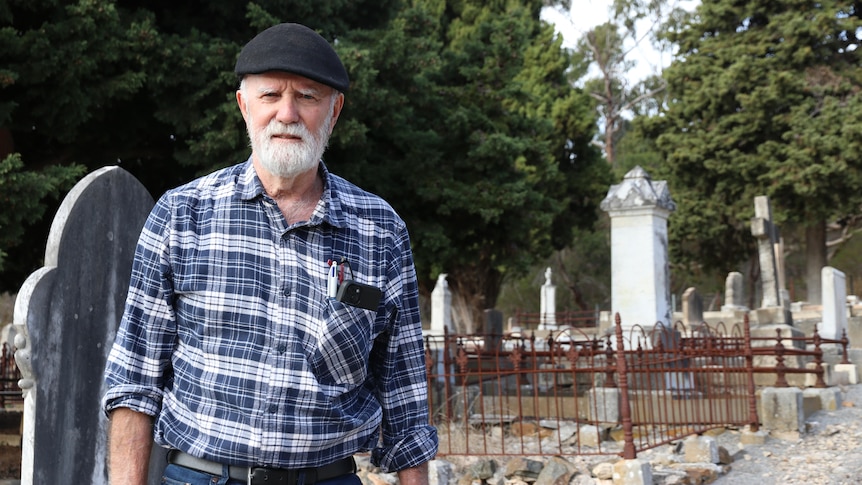 Older man in flat cap and blue check shirt stands in graveyard