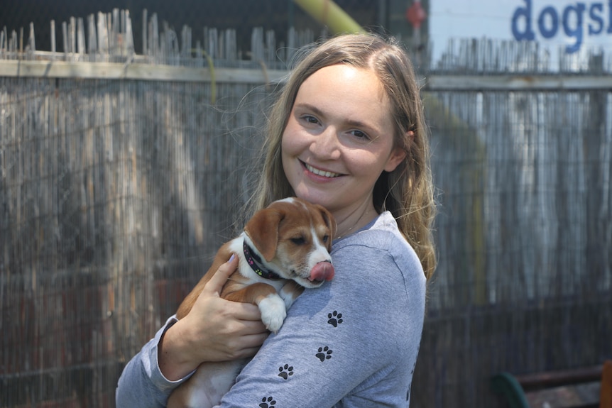 A young blonde woman stands posing for a photo smiling while holding a puppy dog.