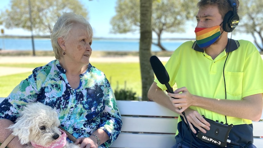 Young man interviewing woman, both wearing masks, the man has a gay pride mask, the woman has a dog on her lap