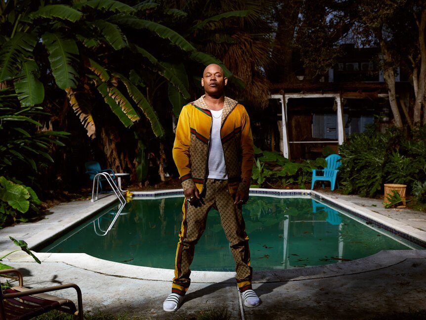Man wearing head-to-toe Gucci standing square to camera, holding its gaze, in front of pool in a backyard.