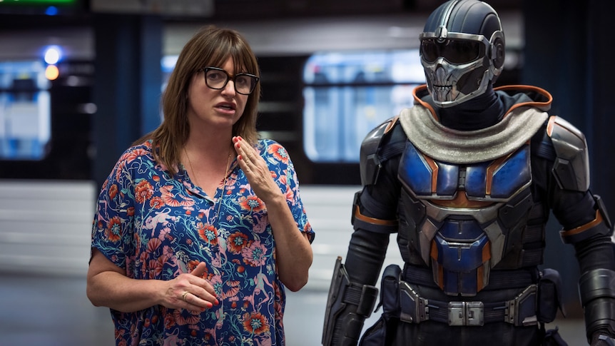 A woman in her early 50s in floral top with glasses directs someone in soldier/robot outfit