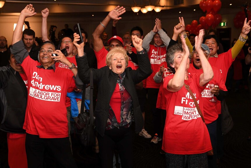 People in red 'Daniel Andrews' shirts cheer at a function centre.