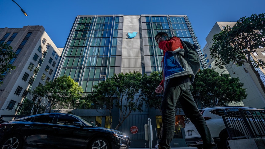 A person talks on the phone while walking past a building with the Twitter logo on the outside