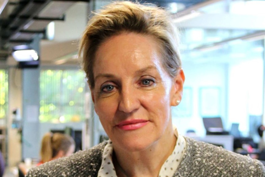 Middle-aged Caucasian woman with blonde her wearing a polka dot shirt and tweed jacket, standing in a newsroom office