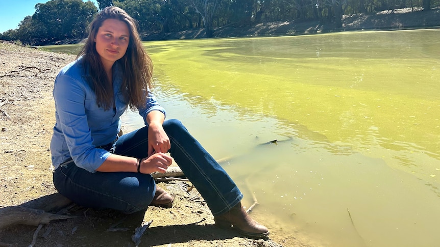 A young, dark-haired woman in a shirt, jeans and boots sits on a fallen branch on the edge of a river.