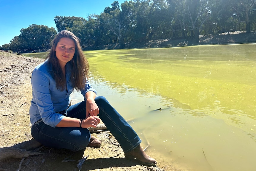 A woman in a blue shirt and blue jeans sits next the Darling River. 