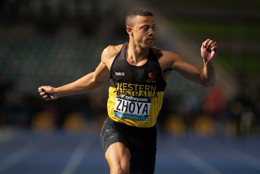 Sashe Zhoya runs wearing a black and yellow singlet, with his arms bent and raised whilst looking to his left