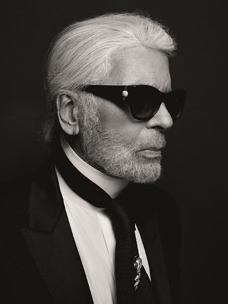 A black-and-white portrait photograph shows Karl Lagerfeld wearing a crisp black suit and tie and dark glasses