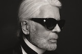 A black-and-white portrait photograph shows Karl Lagerfeld wearing a crisp black suit and tie and dark glasses
