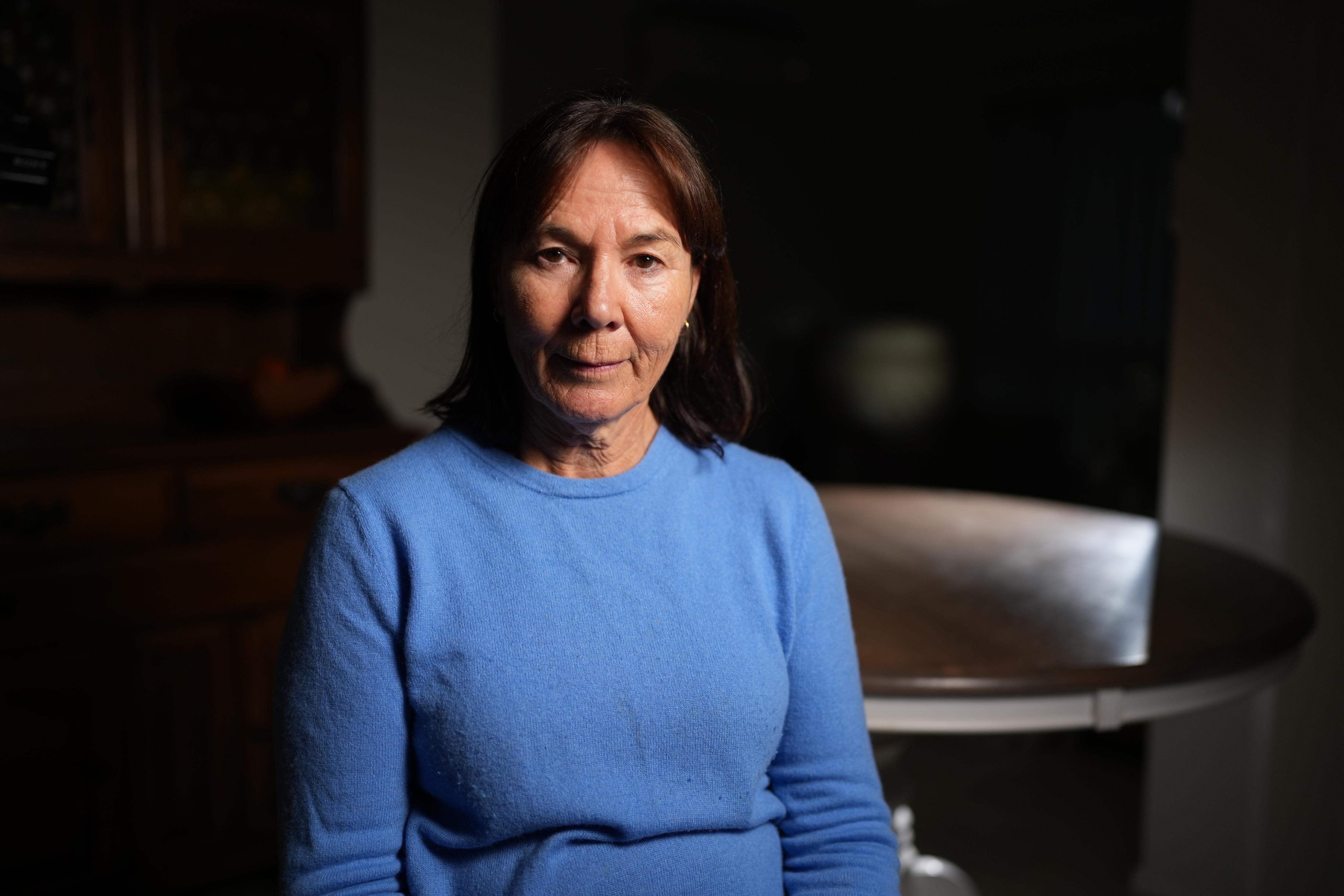 An older woman with shoulder-length dark hair wearing a cornflower blue sweater sits in front of a dining table.