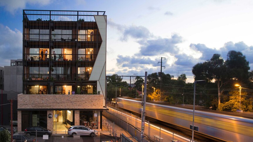 A modern looking apartment building next to a rail line, with people standing on their verandas chatting.