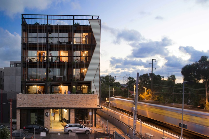 A modern looking apartment building next to a rail line, with people standing on their verandas chatting.