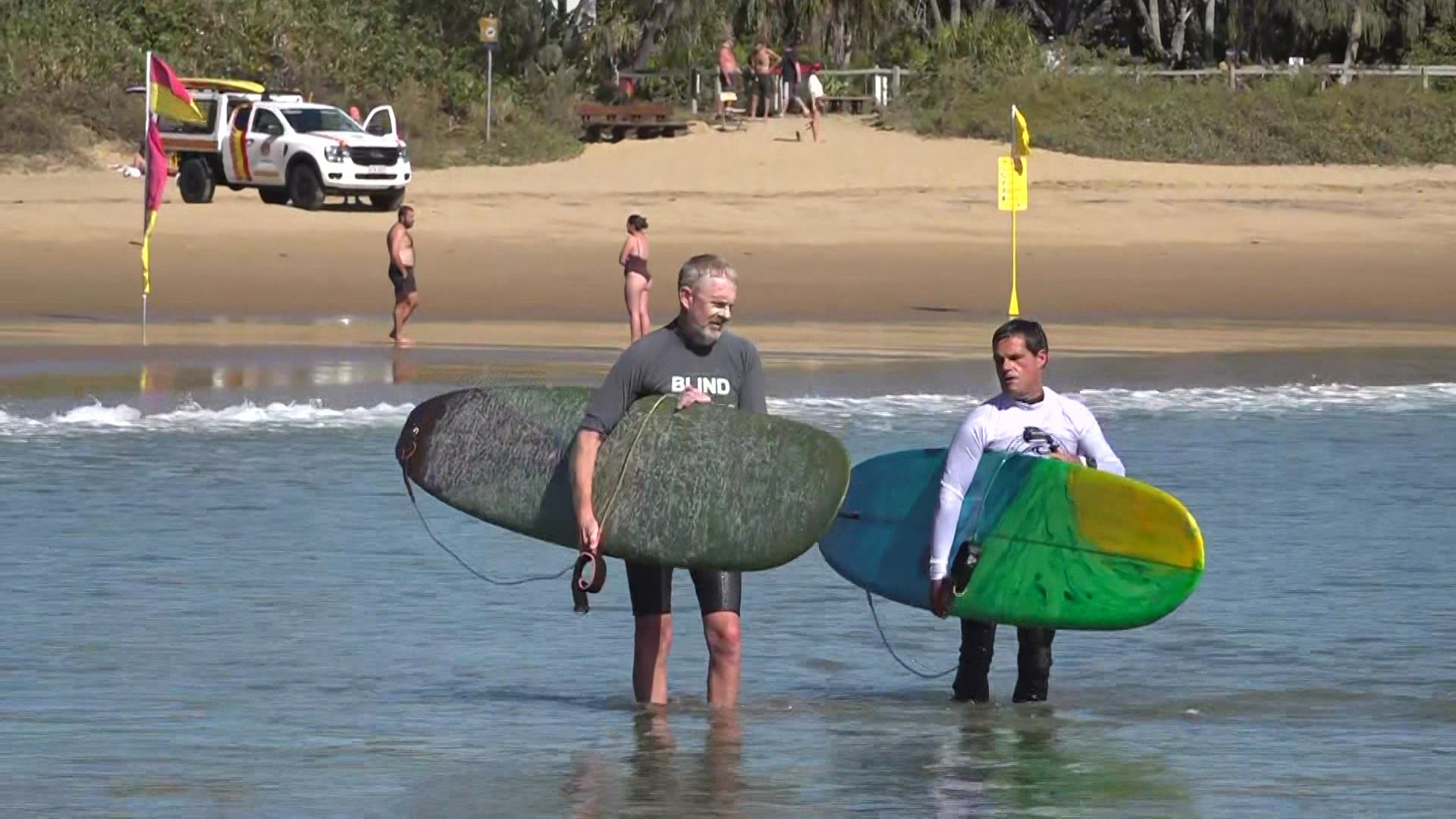 Two men holding surf boards walking into the ocean with the beach in the background