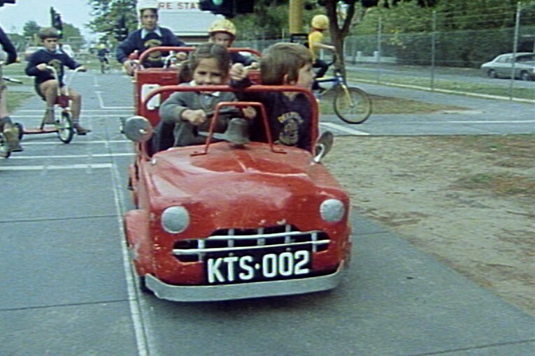 Two children in red miniature car ride ahead of kids on bikes at a miniature road crossing inside a traffic park.