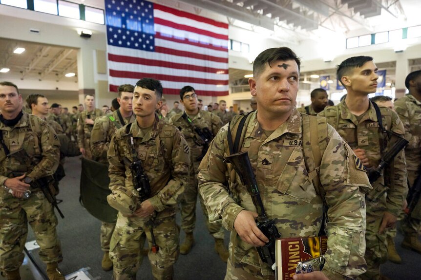 Soldiers stand in front ot US flag, ash on foreheads.