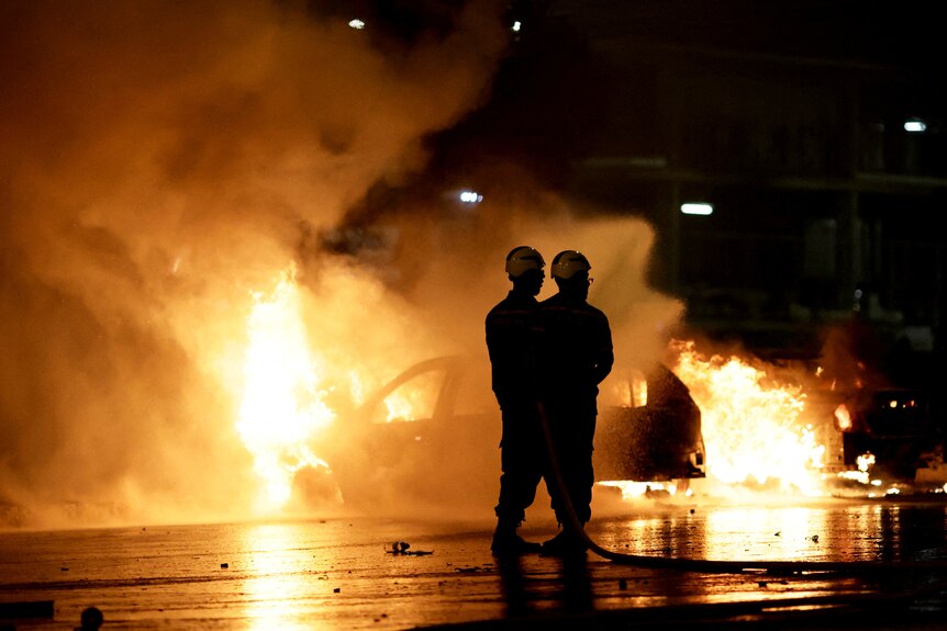Two firemen are standing in front of a car that is fully ablaze