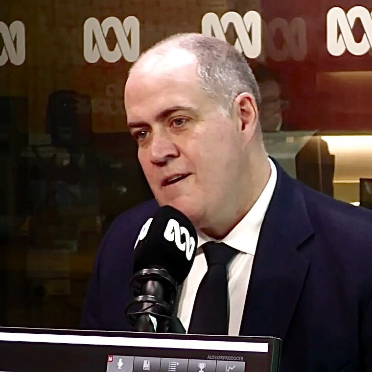A bald man in a suit sits in front of a microphone