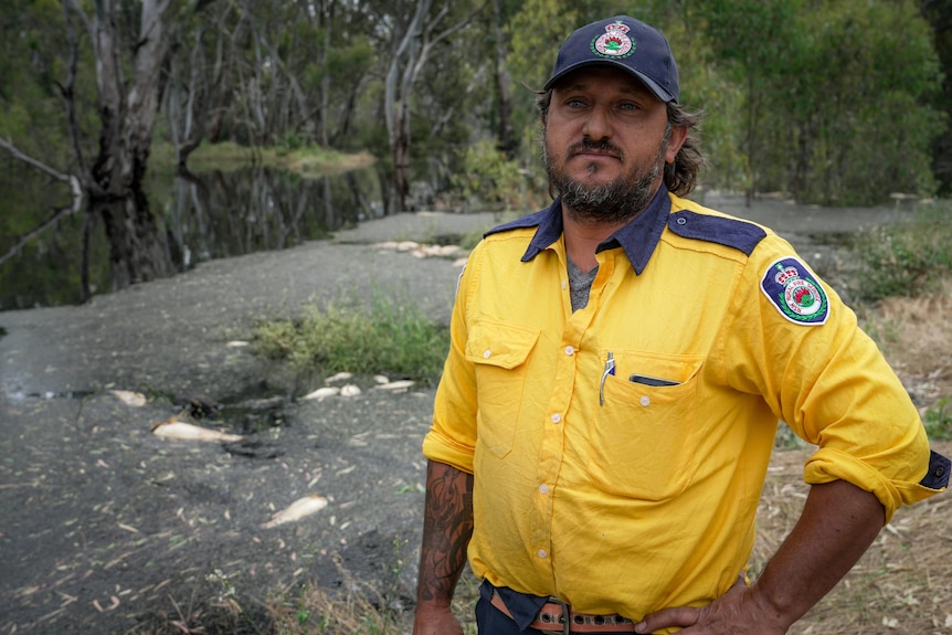 Man in NSW Rural Fire Services uniform standing in front of black river with dead fish floating.