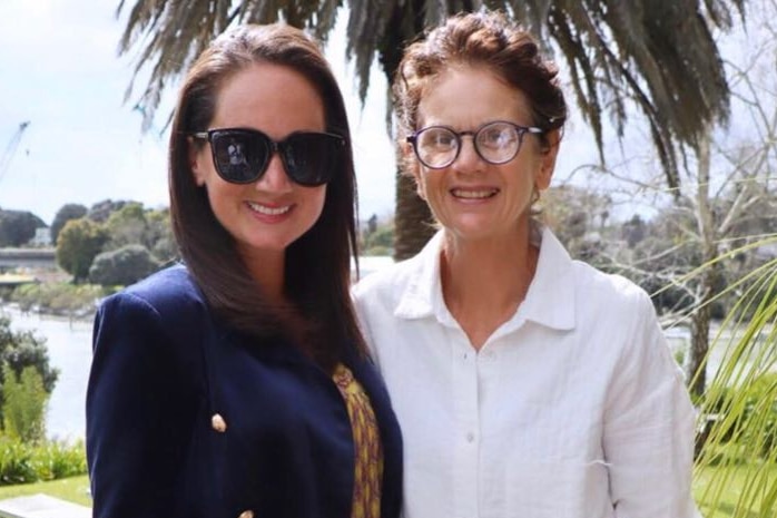 Girl with dark hair and sun glasses wearing a hot pink skirt and blue jacket stands with woman in white shirt and jeans.