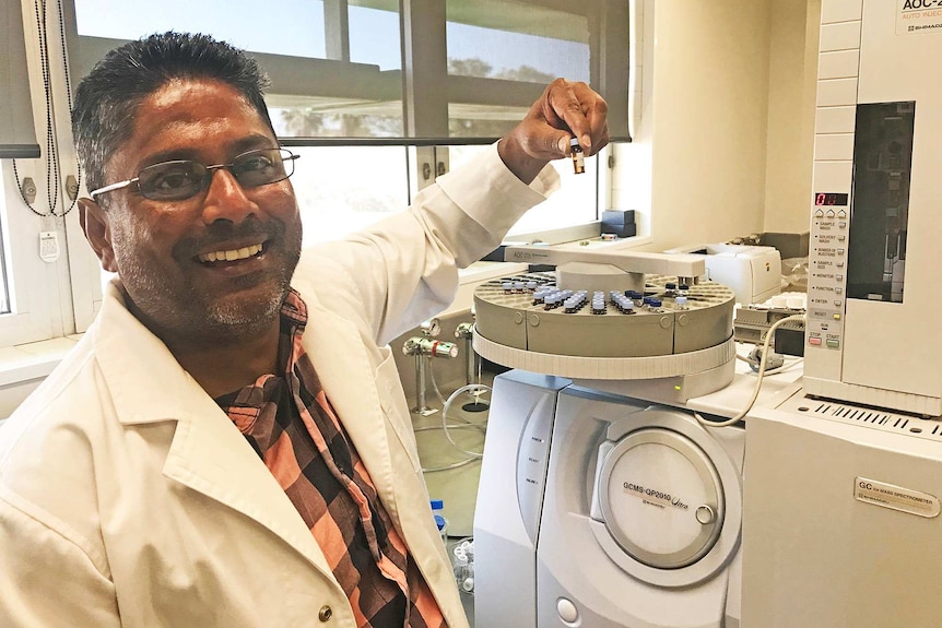 Researcher Dr Mani Naiker wearing glasses and a white lab coat in a lab, holding a sample of red wine.