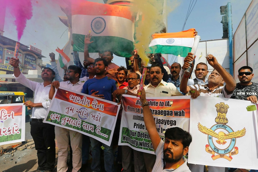 A group of men stand with signs, the Indian flag and flares emitting pink and yellow smoke as they celebrate attack on Kashmir.