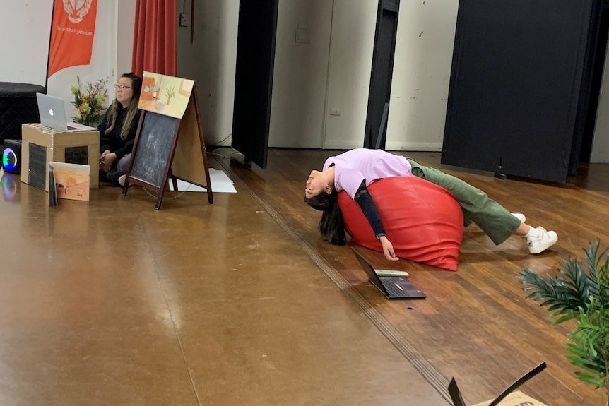 Actor Shelby Brodie lies on her back bored on a red bean bag on a small stage.