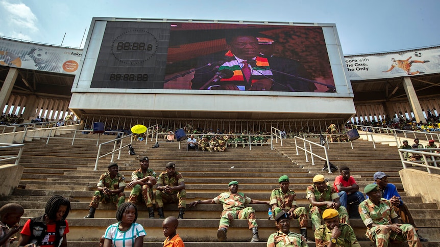 You look up stadium stands sparsely populated to a screen showing the image of Emmerson Mnangagwa.
