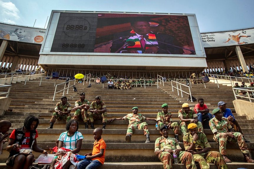 You look up stadium stands sparsely populated to a screen showing the image of Emmerson Mnangagwa.