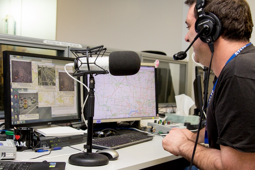 A man smiles while wearing headphones and talking into a microphone, screens showing maps and videos of freeways in front of him