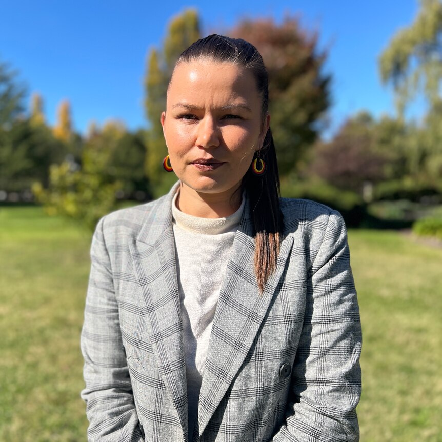 An Indigenous woman in a plaid suit jacket stands in a field looking serious.