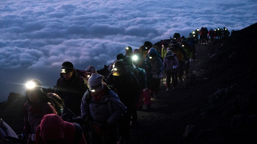Dozens of hikers walk up a steep hill high above the cloud level wearing headlamps