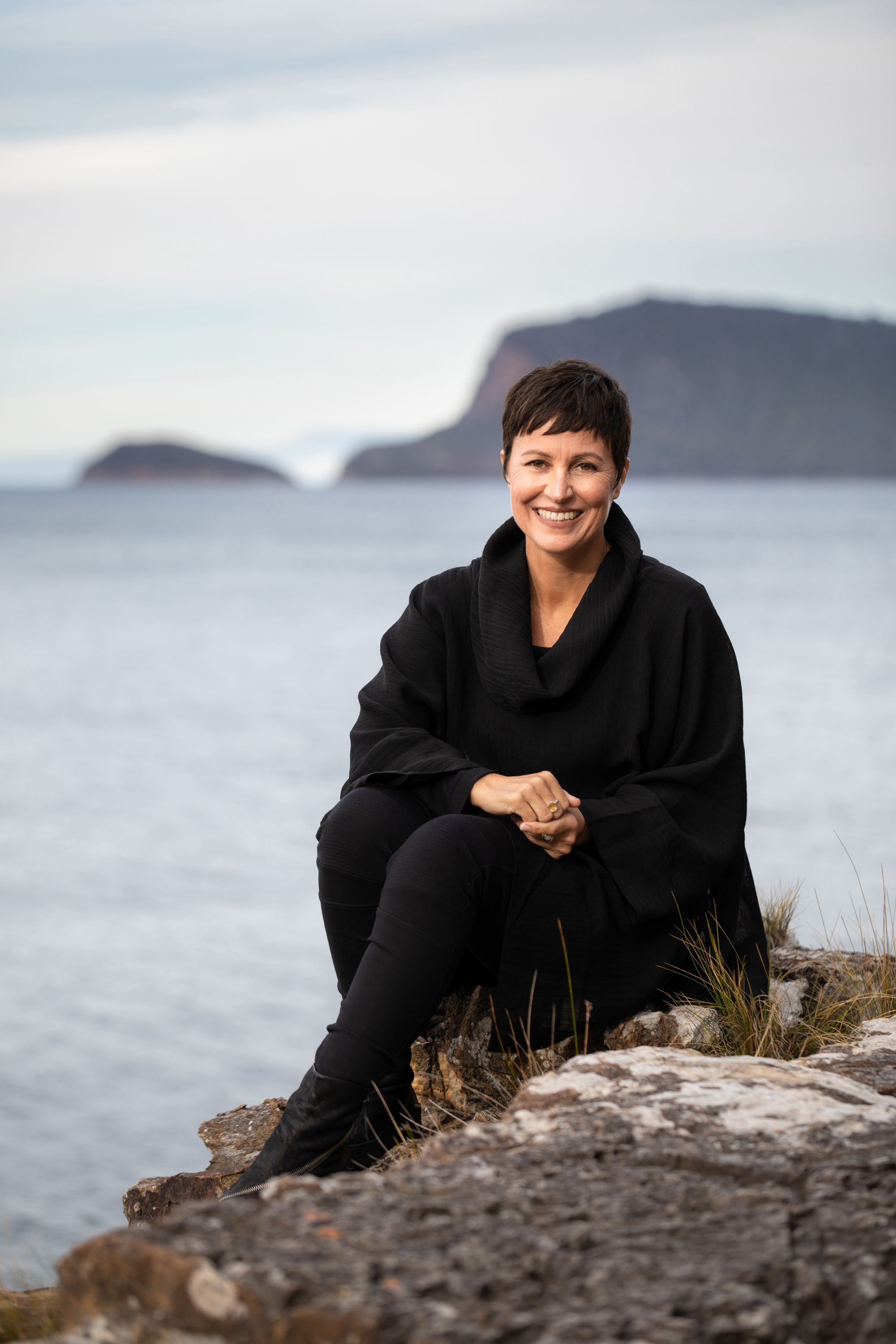 A 50-something brunette woman, with short hair and a fringe, smiles at the camera. She sits on a rock on a coastline.