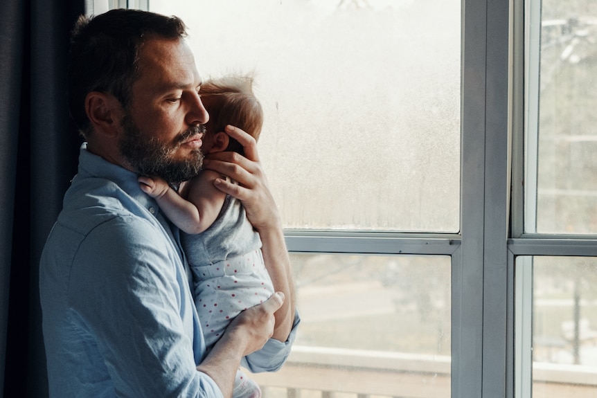 A father holds a baby, as he stands at a window.