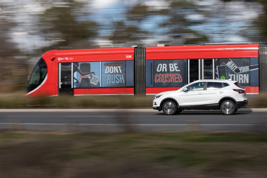 A light rail vehicle travels down the road and a white car drives beside it.