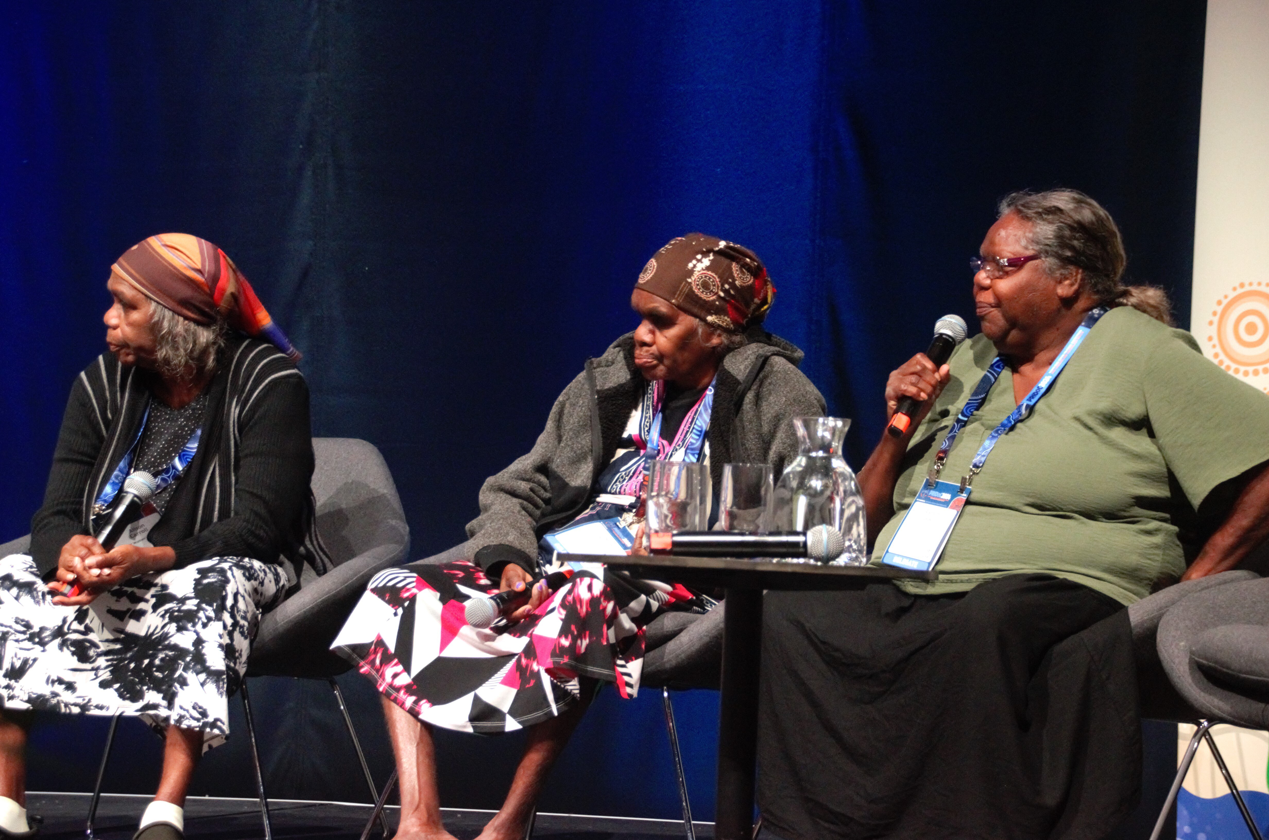 Ngangkari Pantjiti Lewis sits between two other people on a stage, with one holding a microphone