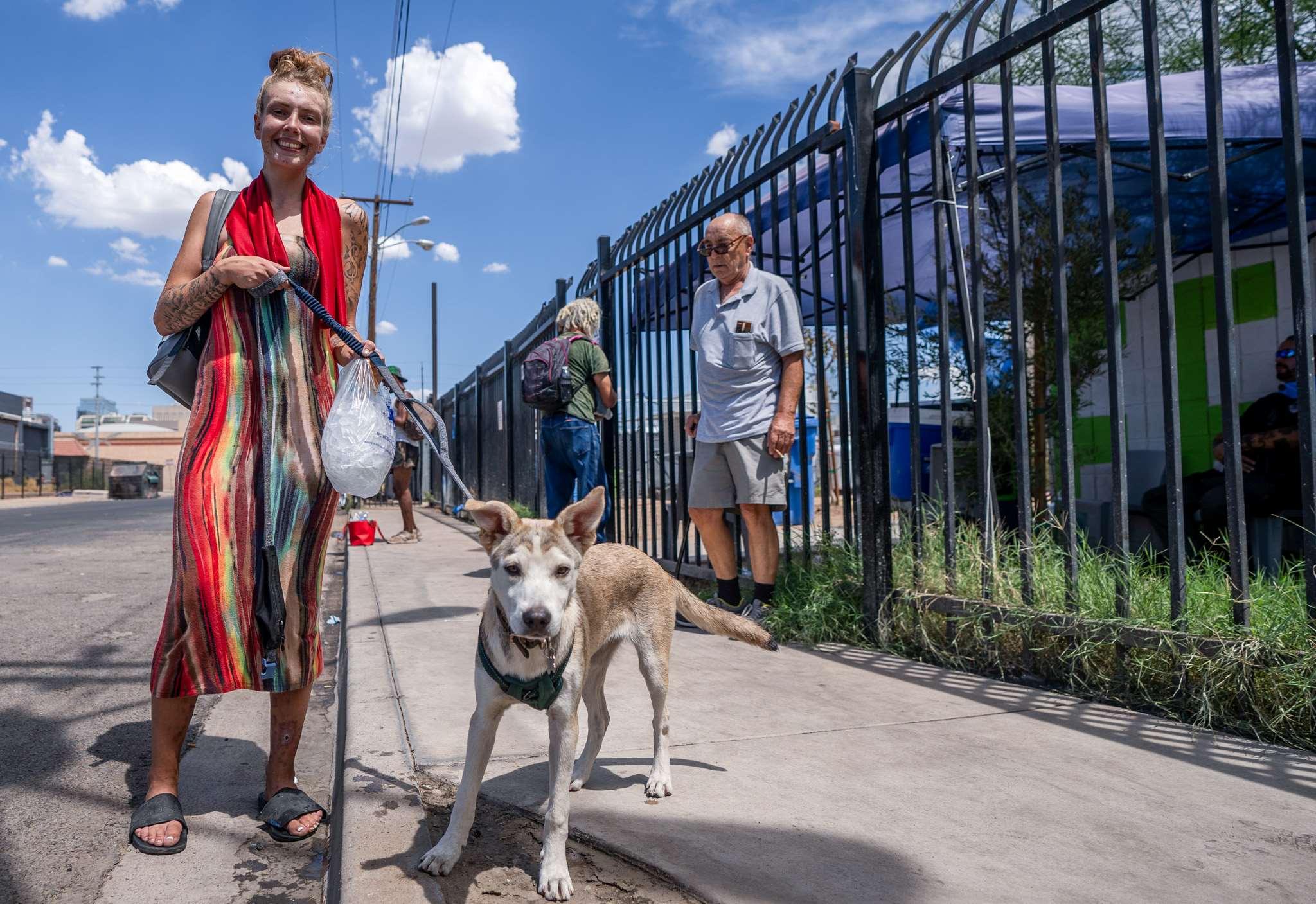 A woman wearing a colourful dress stands on a street holding a bag of ice and her dog on a leash.
