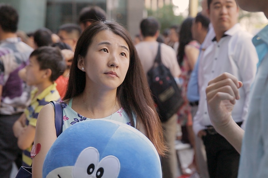 Colour still of Xu Min in a crowded outdoor area, holding plush toy and looking up in 2019 documentary Leftover Women.