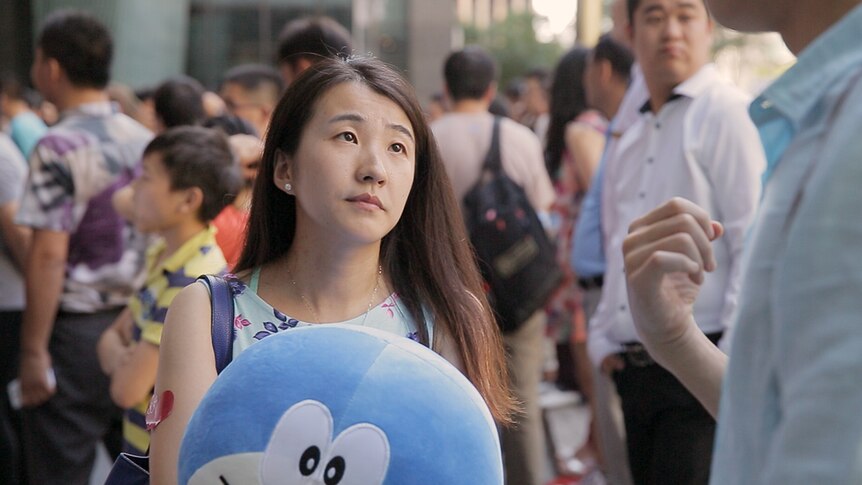 Colour still of Xu Min in a crowded outdoor area, holding plush toy and looking up in 2019 documentary Leftover Women.