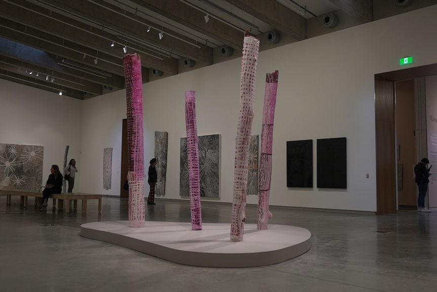 Four bright pink poles amde from tree trunks are standing on a white platform with three bark paintings behind them