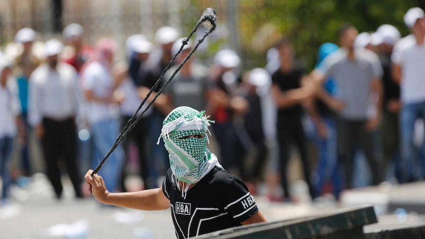 A man with his face covered swings a slingshot as a crowd of people stand in the background.