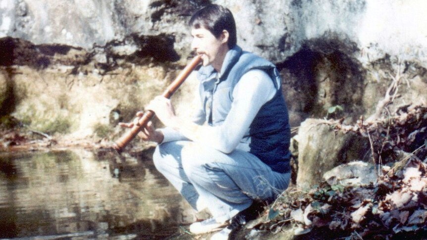 A man plays the shakuhachi while crouching on a rock in front of a body of water.