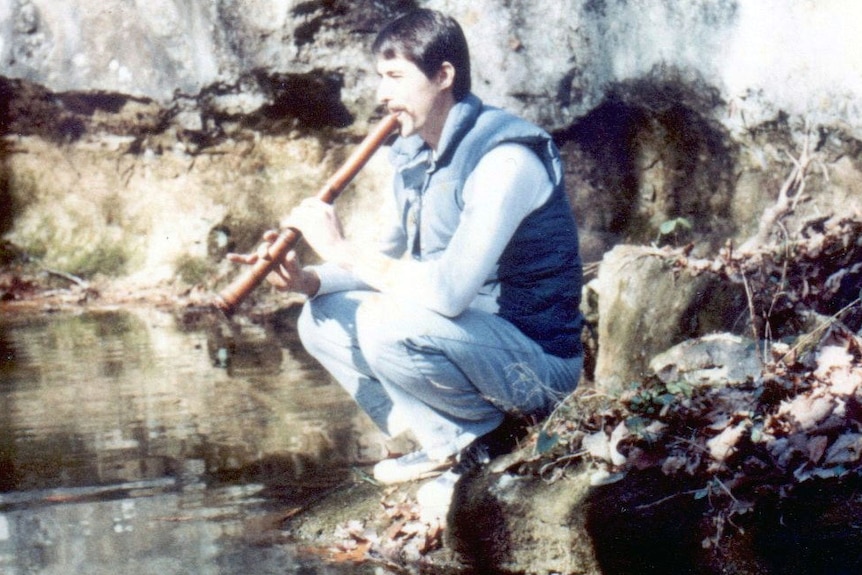 A man plays the shakuhachi while crouching on a rock in front of a body of water.