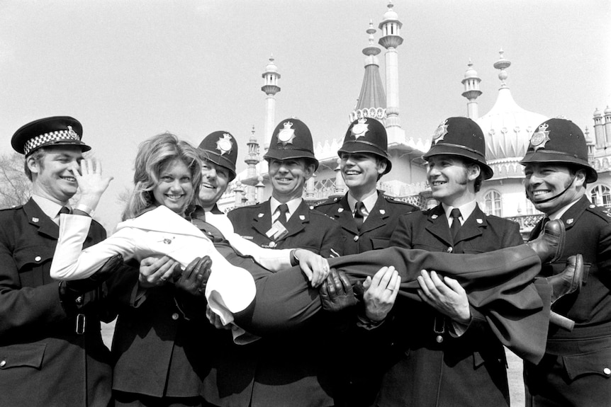 Olivia Newton John waves as she is held up by a row of six policeman in Brighton, UK