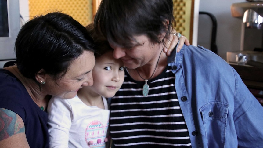 Claudia Alessi and Zoe Goss both hug their daughter Zara in the lounge room of their house.