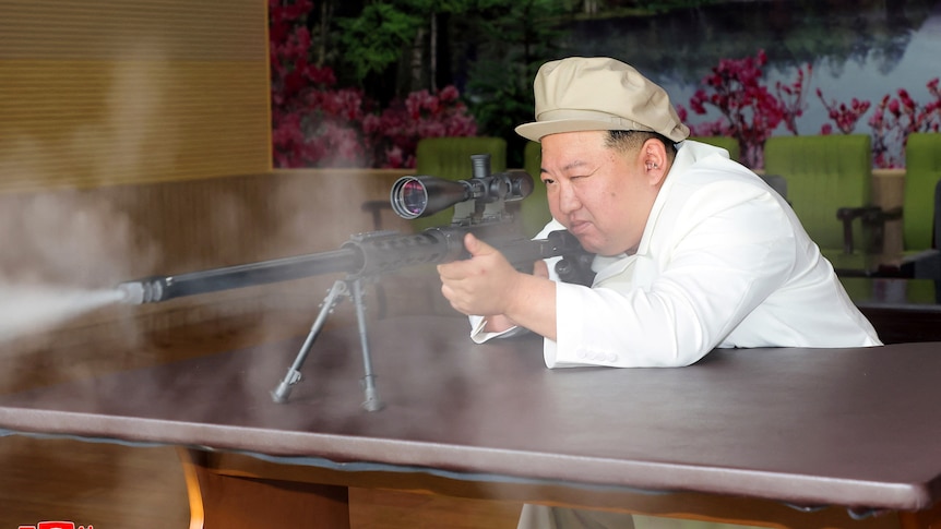 Kim Jong Un leans over a table, looking through a magnifier as he points a gun.