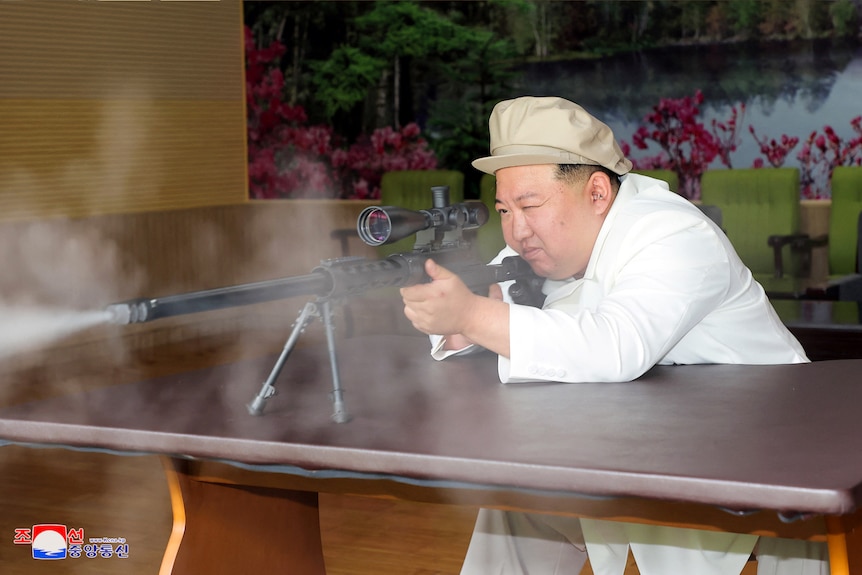 Kim Jong Un leans over a table, looking through a magnifier as he points a gun.