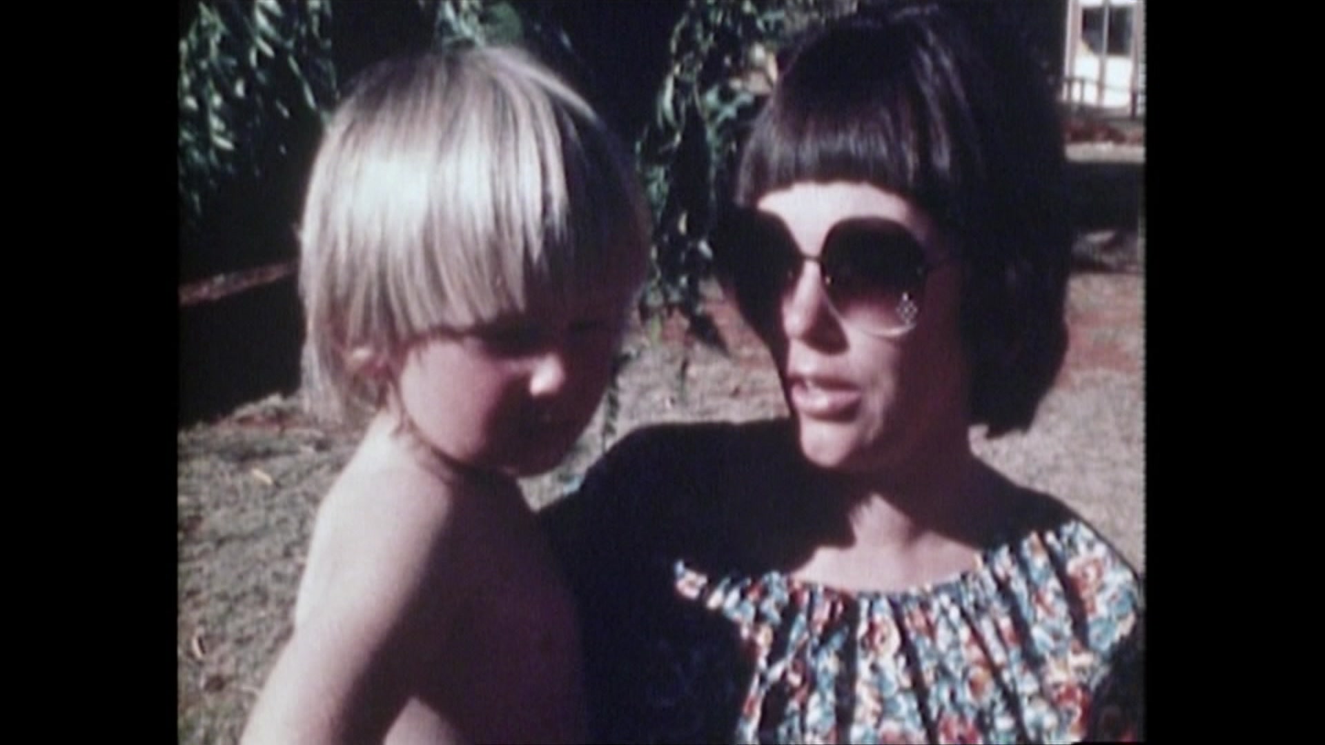 An archival still from a video shows a mother with short dark hair, holding her young son as she speaks to a journalist.