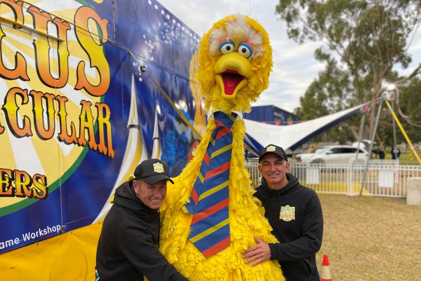 Two men wearing black hug a giant yellow Big Bird character outside a circus tent